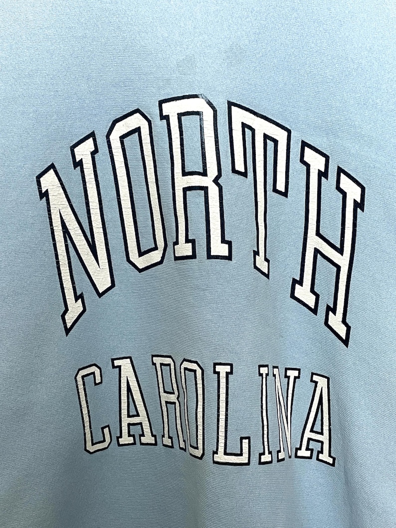 K&ouml;nnte beinhalten: Hellblauer Pullover mit wei&szlig;er und schwarzer Schrift, die "NORTH CAROLINA" lautet.