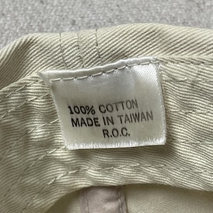 May include: A beige cotton fabric interior of a cap featuring a white rectangular tag with black text that reads 100% COTTON, MADE IN TAIWAN, R.O.C. The fabric shows visible stitching lines along the brim and crown area of the headwear item.