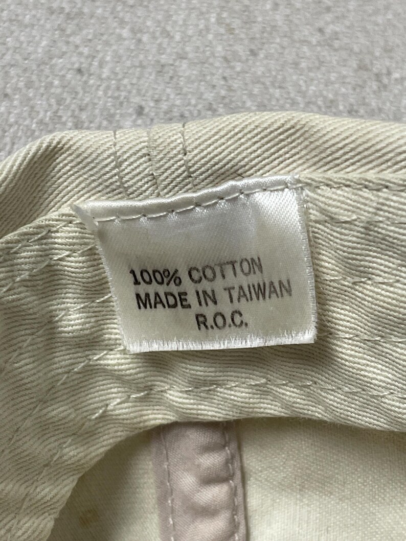 May include: A beige cotton fabric interior of a cap featuring a white rectangular tag with black text that reads 100% COTTON, MADE IN TAIWAN, R.O.C. The fabric shows visible stitching lines along the brim and crown area of the headwear item.