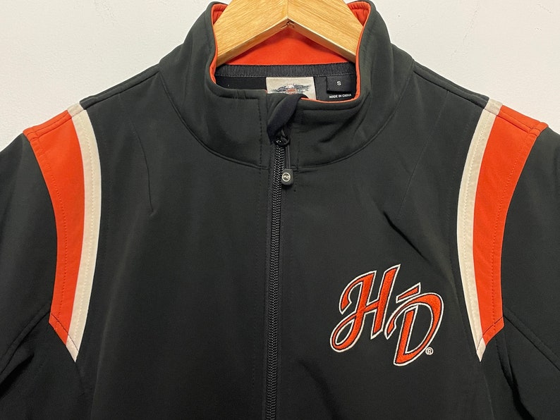May include: Black zippered jacket with orange and white stripes on the sleeves. The jacket has a large "HD" logo on the front.