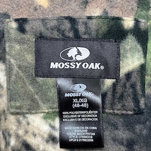May include: A close-up of a tag from a Mossy Oak hunting garment. The tag is black with white text and the Mossy Oak logo. The tag reads XL/XG (46-48) and includes fabric and care information. The garment has a camouflage pattern.