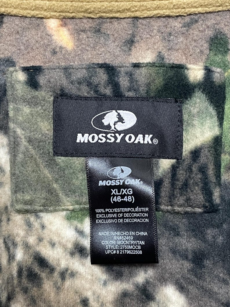 May include: A close-up of a tag from a Mossy Oak hunting garment. The tag is black with white text and the Mossy Oak logo. The tag reads XL/XG (46-48) and includes fabric and care information. The garment has a camouflage pattern.