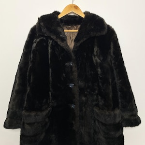 Puede incluir: Un abrigo de piel sintética marrón oscuro con cuello, cierre de botones y dos bolsillos delanteros. El abrigo está colgado de una percha de madera sobre un fondo blanco. El abrigo parece ser vintage.