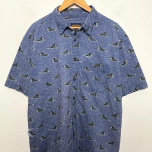Op de afbeelding: Een overhemd met korte mouwen en knoopsluiting van gemêleerde blauwe stof met een all-over print van donkergekleurde honden. Het overhemd heeft een kraag, een enkele borstzak en een label aan de binnenkant van de nek met de tekst Woolrich.