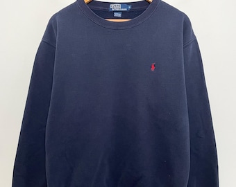 May include: Navy blue sweatshirt with a crew neck and long sleeves. A small red logo is embroidered on the left chest. The sweatshirt is hanging on a wooden hanger against a white background.