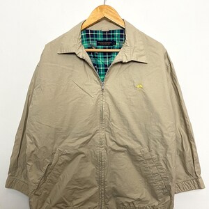 May include: A beige zip-up jacket with a green and black plaid lining. The jacket has a small embroidered logo on the left chest.