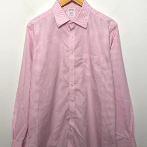 Pode incluir: Camisa xadrez rosa e branca com mangas compridas, punhos abotoados e bolso no peito. A camisa está pendurada em um cabide de madeira em um fundo branco. A camisa tem uma gola clássica.