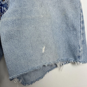 May include: Light blue denim shorts with a distressed, frayed hem. The shorts show signs of wear with a small tear and discoloration, suggesting a vintage or well-loved item. The fabric has a classic denim texture.