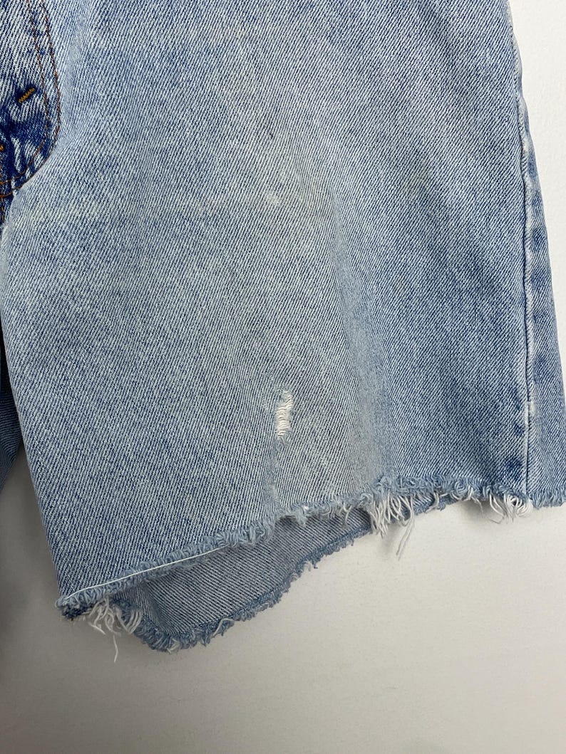 May include: Light blue denim shorts with a distressed, frayed hem. The shorts show signs of wear with a small tear and discoloration, suggesting a vintage or well-loved item. The fabric has a classic denim texture.