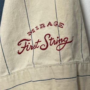 May include: A cream-colored short-sleeved shirt featuring thin vertical navy blue pinstripes. The fabric is embroidered with the text "MIRAGE" in small red letters above the larger red cursive script text "First String" on the sleeve.