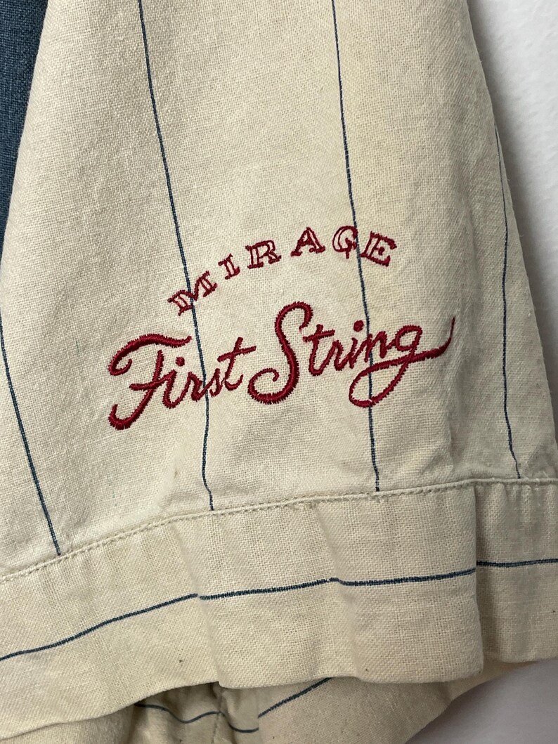 May include: A cream-colored short-sleeved shirt featuring thin vertical navy blue pinstripes. The fabric is embroidered with the text "MIRAGE" in small red letters above the larger red cursive script text "First String" on the sleeve.