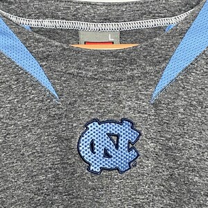 Puede incluir: Camiseta deportiva gris con detalles en azul claro y un logotipo bordado en azul claro. La camiseta tiene cuello redondo y una etiqueta con la talla "L". El logotipo es un emblema estilizado.
