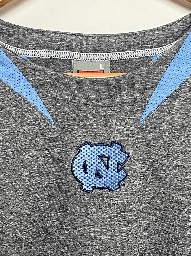 Puede incluir: Camiseta deportiva gris con detalles en azul claro y un logotipo bordado en azul claro. La camiseta tiene cuello redondo y una etiqueta con la talla "L". El logotipo es un emblema estilizado.
