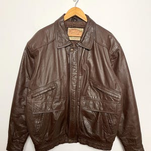 May include: A brown leather jacket with a collared neckline and two front pockets. The jacket is hanging on a wooden hanger against a white background. The jacket has a button-up closure and a label on the collar.