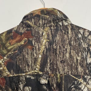 Pu&ograve; includere: Giacca da caccia mimetica con abbottonatura e colletto. La giacca presenta un motivo marrone, verde e beige, che ricorda un ambiente forestale. La giacca &egrave; appesa a un muro bianco.
