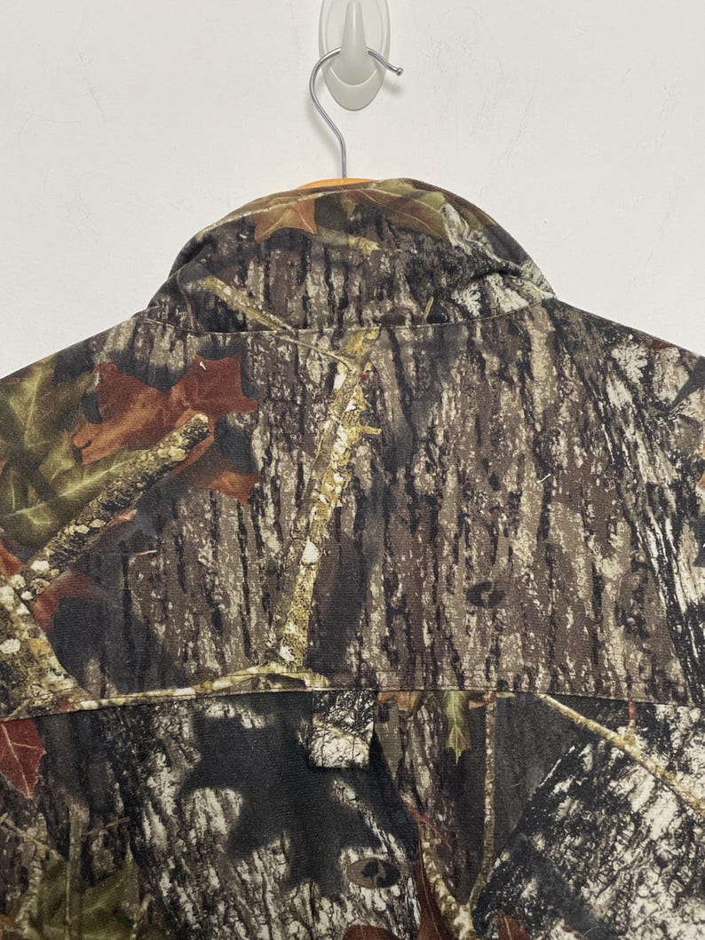 Pu&ograve; includere: Giacca da caccia mimetica con abbottonatura e colletto. La giacca presenta un motivo marrone, verde e beige, che ricorda un ambiente forestale. La giacca &egrave; appesa a un muro bianco.