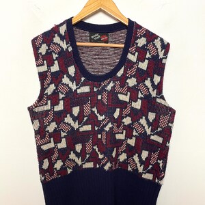 Pode incluir: Um colete de malha sem mangas com um padrão geométrico em azul-marinho, vermelho escuro e branco sujo. A peça tem uma bainha canelada em azul-marinho sólido e um decote redondo canelado a condizer. Uma etiqueta interior diz "gruff stuff".