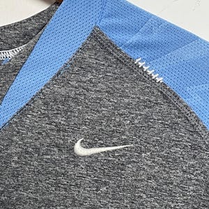 Puede incluir: Primer plano de una camiseta deportiva gris con un panel de malla azul claro en el escote. Un logotipo blanco de Nike swoosh es visible en el pecho. La camiseta est&aacute; colgada en una percha de madera.