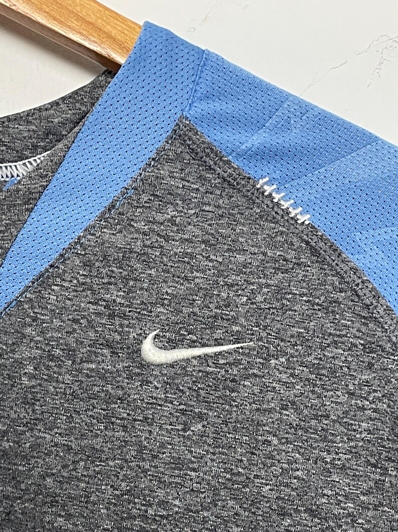 Puede incluir: Primer plano de una camiseta deportiva gris con un panel de malla azul claro en el escote. Un logotipo blanco de Nike swoosh es visible en el pecho. La camiseta est&aacute; colgada en una percha de madera.