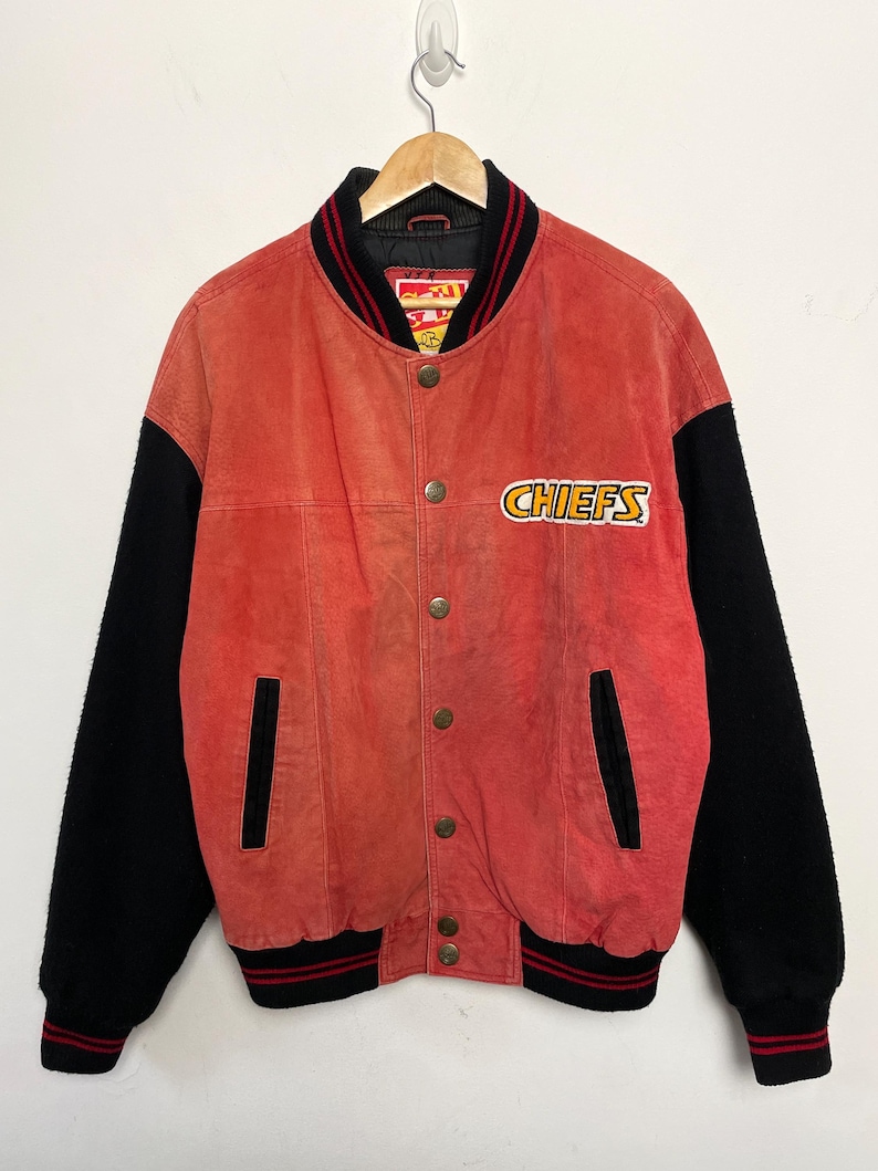 Puede incluir: Una chaqueta de b&eacute;isbol de gamuza roja y lana negra con un logotipo de Chiefs en la parte delantera. La chaqueta tiene un cuello y pu&ntilde;os a rayas rojas y negras.