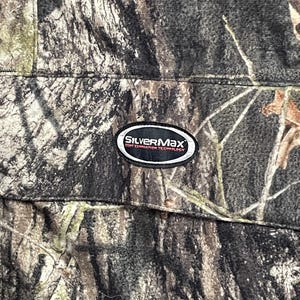 Pode incluir: Close-up de tecido camuflado com um padr&atilde;o marrom, verde e cinza. O tecido tem uma costura e um patch SilverMax em preto e branco com texto. O design de camuflagem se assemelha a uma floresta.