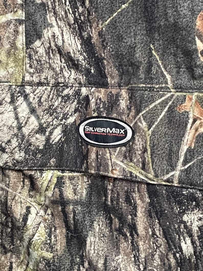 Pode incluir: Close-up de tecido camuflado com um padr&atilde;o marrom, verde e cinza. O tecido tem uma costura e um patch SilverMax em preto e branco com texto. O design de camuflagem se assemelha a uma floresta.