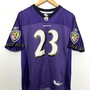 May include: Purple football jersey with the number 23 in gold and white. The jersey has the Baltimore Ravens logo on the left and right sleeves. The jersey is on a hanger.