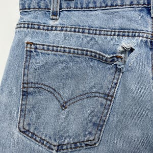 May include: Close-up of light blue denim jeans with a distressed pocket. The jeans feature a classic five-pocket design with a worn, ripped pocket. The Levi's brand logo is visible on the back pocket. The jeans are hanging on a metal hanger.