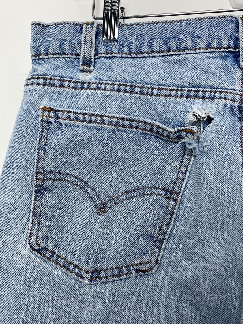 May include: Close-up of light blue denim jeans with a distressed pocket. The jeans feature a classic five-pocket design with a worn, ripped pocket. The Levi's brand logo is visible on the back pocket. The jeans are hanging on a metal hanger.