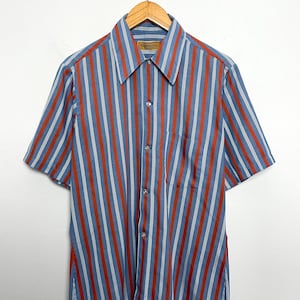 Op de afbeelding: Korte mouwen overhemd met verticale strepen in blauw, rood en wit. Het overhemd heeft een kraag, een borstzak en hangt aan een houten hanger.