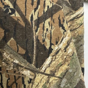 May include: Close-up of a camouflage pattern fabric. The design features a blend of brown, tan, and black shapes, mimicking a forest environment. The fabric appears to be a medium-weight material, suitable for outdoor apparel.