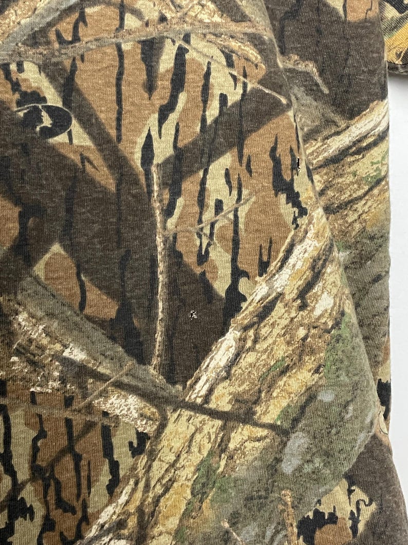 May include: Close-up of a camouflage pattern fabric. The design features a blend of brown, tan, and black shapes, mimicking a forest environment. The fabric appears to be a medium-weight material, suitable for outdoor apparel.