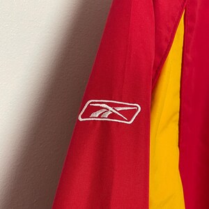 Puede incluir: Primer plano de una manga de chaqueta roja y amarilla. La chaqueta presenta un logotipo blanco de Reebok sobre la tela roja. La manga tiene un panel amarillo en el lateral.