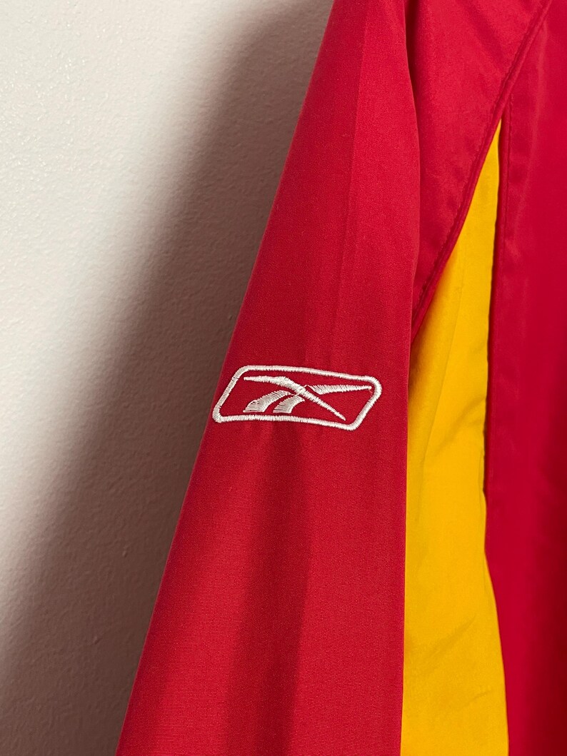 Puede incluir: Primer plano de una manga de chaqueta roja y amarilla. La chaqueta presenta un logotipo blanco de Reebok sobre la tela roja. La manga tiene un panel amarillo en el lateral.