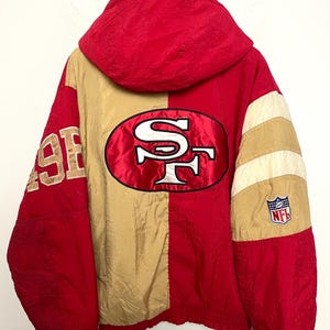 May include: A red and gold hooded jacket featuring the San Francisco 49ers logo prominently on the back. The jacket includes the text 49ERS on the right sleeve and an NFL shield patch on the left sleeve, designed with a color-blocked pattern.