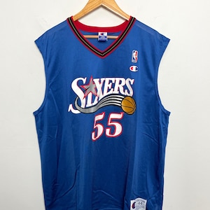 Può includere: Una canotta da basket blu vintage con la scritta "Sixers" in bianco e rosso, insieme al numero "55" in rosso. La canotta ha un bordo rosso e nero intorno al colletto e alle ascelle. È visibile un logo Champion.