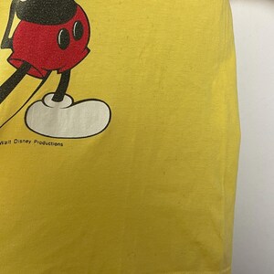 K&ouml;nnte beinhalten: Gelbes T-Shirt mit einem Teilbild von Mickey Mouse. Die Zeichentrickfigur ist in Schwarz, Rot und Wei&szlig; dargestellt. Der Text "Walt Disney Productions" ist sichtbar. Das T-Shirt hat eine einfarbige gelbe Farbe.