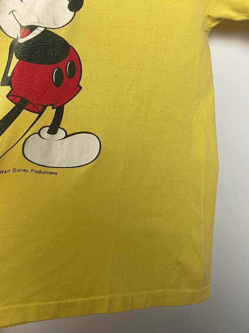 K&ouml;nnte beinhalten: Gelbes T-Shirt mit einem Teilbild von Mickey Mouse. Die Zeichentrickfigur ist in Schwarz, Rot und Wei&szlig; dargestellt. Der Text "Walt Disney Productions" ist sichtbar. Das T-Shirt hat eine einfarbige gelbe Farbe.