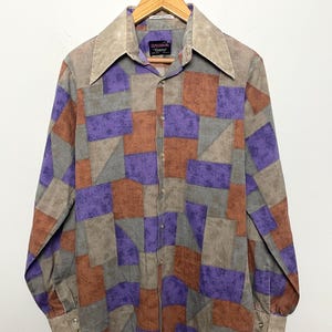 May include: A vintage patchwork shirt with a wide collar, button-down front, and long sleeves. The shirt features a geometric pattern of brown, purple, gray, and tan squares and triangles. The shirt is hanging on a wooden hanger.