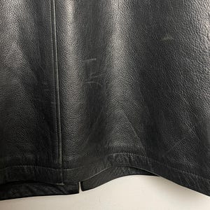 May include: Close-up of the bottom edge of a black leather jacket. The jacket has a seam down the center back and a curved hem.