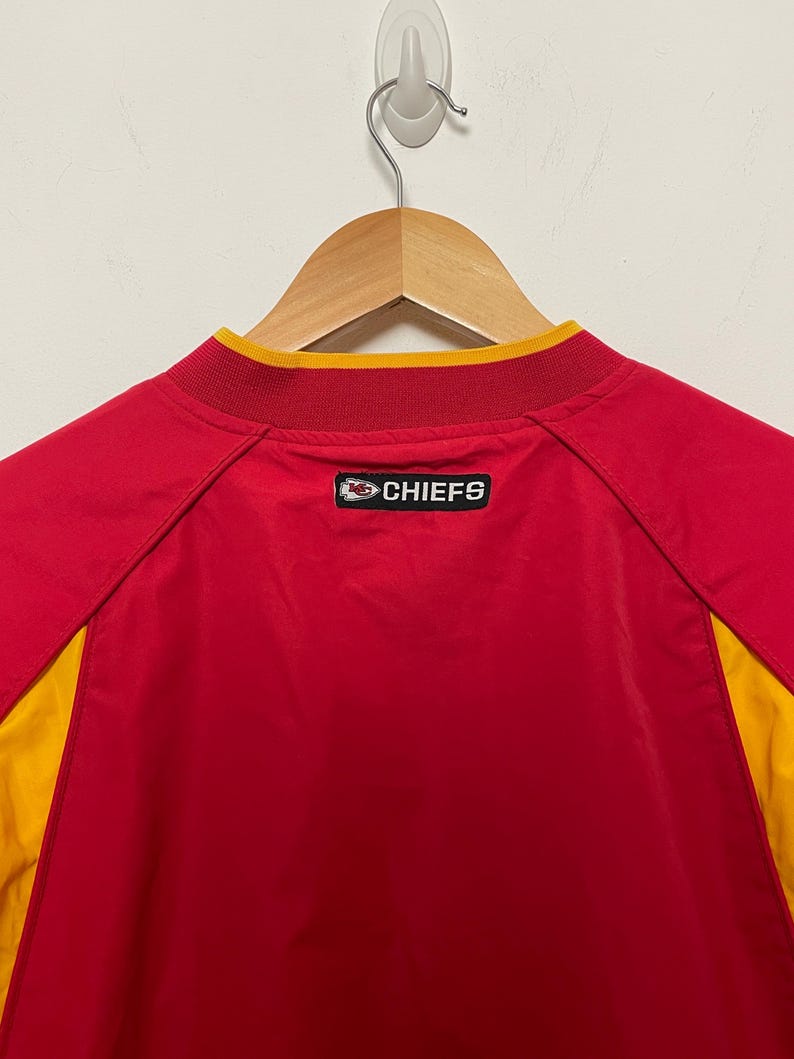 Puede incluir: Chaqueta deportiva roja y amarilla con la palabra "CHIEFS" en la espalda. La chaqueta est&aacute; colgada de una percha de madera. El cuello es amarillo y la chaqueta es roja con paneles laterales amarillos.
