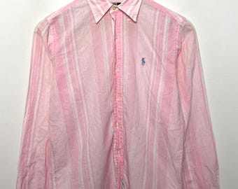 May include: Pink and white striped button-down shirt with long sleeves. The shirt features a small blue logo on the chest. The shirt is hanging on a wooden hanger against a white background. The shirt is made of a lightweight fabric.