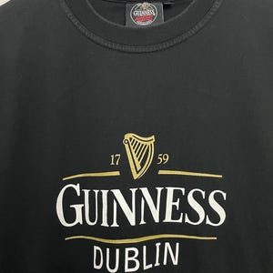 May include: Black t-shirt featuring the Guinness logo in gold and white. The logo includes a harp, the numbers 17 and 59, and the word "Dublin" beneath the Guinness name.