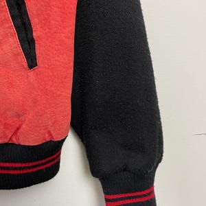 Puede incluir: Una chaqueta de b&eacute;isbol roja y negra con ribetes de rayas rojas y blancas en los pu&ntilde;os y la cintura.