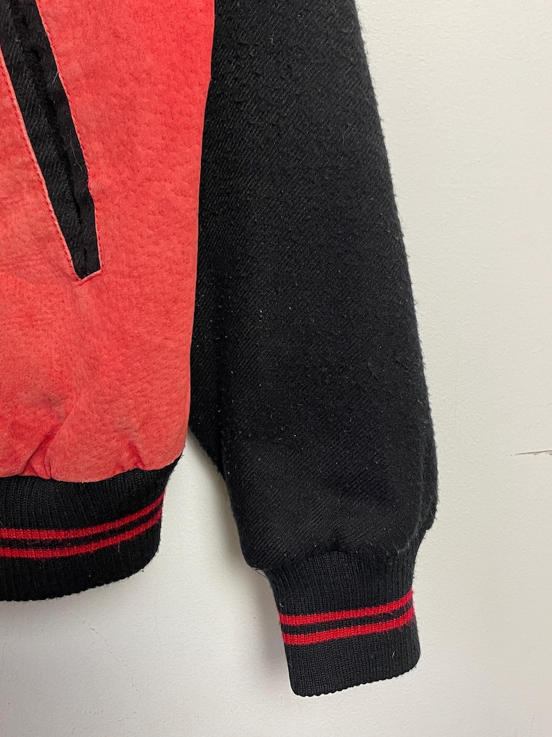 Puede incluir: Una chaqueta de b&eacute;isbol roja y negra con ribetes de rayas rojas y blancas en los pu&ntilde;os y la cintura.