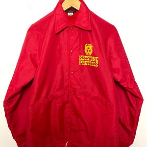 May include: A bright red nylon coach jacket featuring a classic collar, long sleeves with ribbed cuffs, and a snap-button front closure. The left chest displays a yellow embroidered logo with the text Keystone Pretzels above a pretzel graphic.