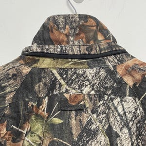 Pode incluir: Close-up da gola de um casaco camuflado com z&iacute;per preto. O casaco apresenta um padr&atilde;o castanho, verde e preto, que se assemelha a um ambiente florestal. O casaco est&aacute; pendurado numa parede branca.