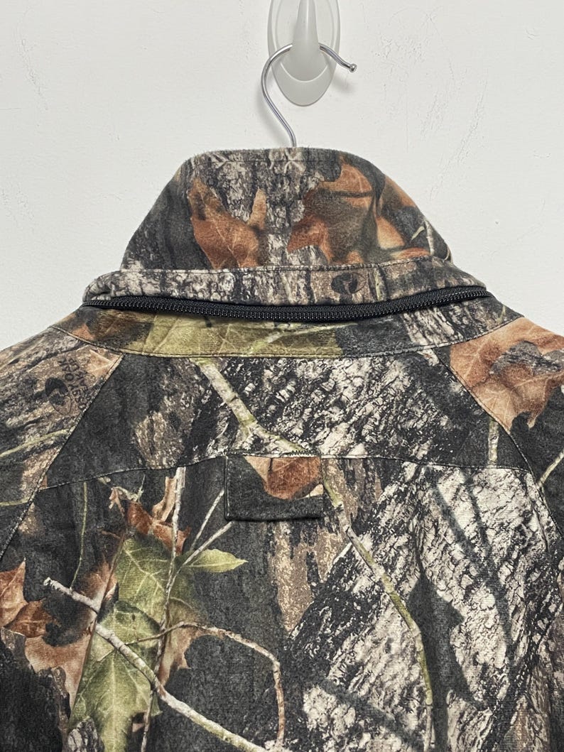 Pode incluir: Close-up da gola de um casaco camuflado com z&iacute;per preto. O casaco apresenta um padr&atilde;o castanho, verde e preto, que se assemelha a um ambiente florestal. O casaco est&aacute; pendurado numa parede branca.