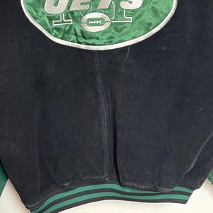 Pode incluir: Casaco vintage preto e verde com o logotipo dos New York Jets. O logotipo est&aacute; em um oval verde com borda branca, com letras prateadas e um gr&aacute;fico de futebol americano. A bainha do casaco &eacute; listrada em verde e preto.