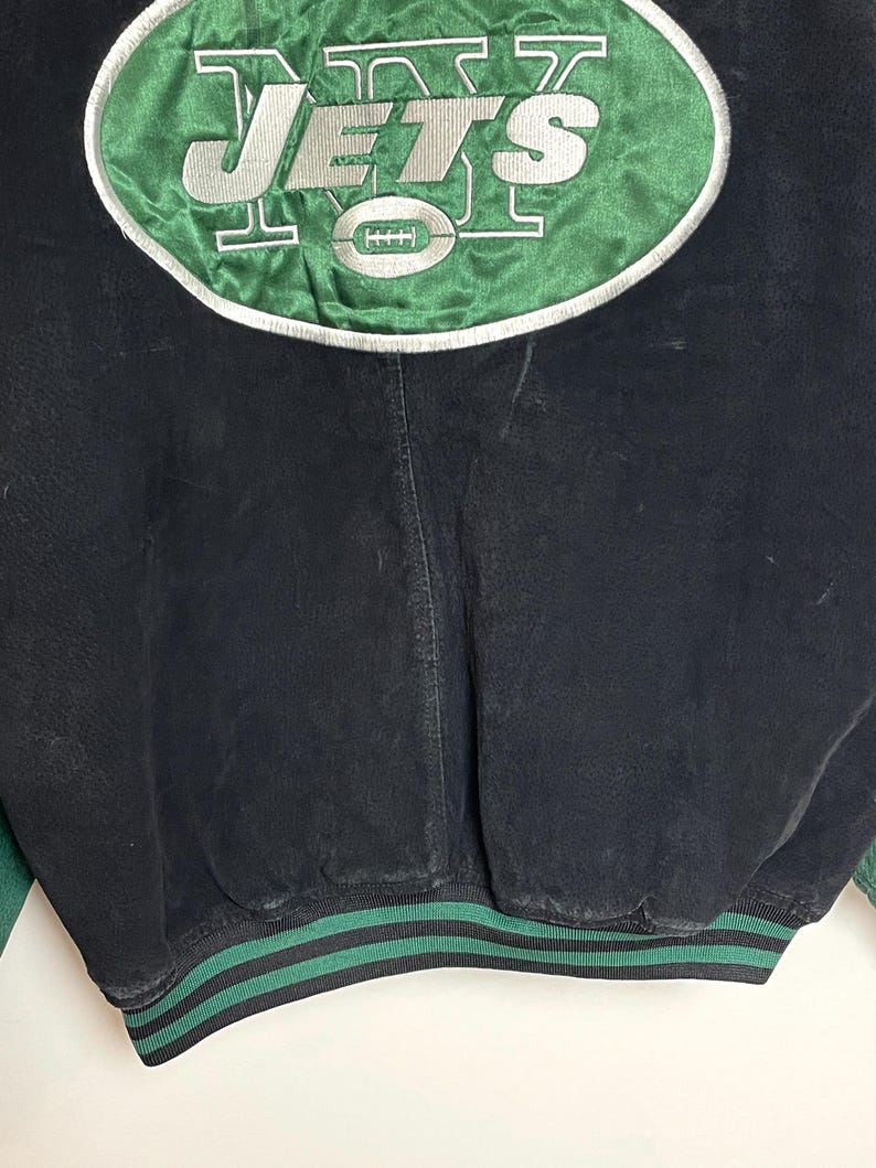 Pode incluir: Casaco vintage preto e verde com o logotipo dos New York Jets. O logotipo est&aacute; em um oval verde com borda branca, com letras prateadas e um gr&aacute;fico de futebol americano. A bainha do casaco &eacute; listrada em verde e preto.